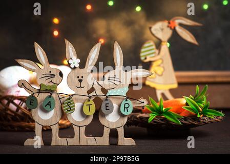 Drei hölzerne Osterhasen mit Ostereiern sind als Dekoration auf einem Holztisch vor einem Fenster mit festlicher Dekoration ausgestellt Stockfoto