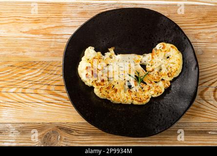 Gebratene Blumenkohlscheiben auf schwarzem Teller, gebackene Blumenkohlsteaks, gebratenes Kohlsteak mit Kräutern, Draufsicht Stockfoto