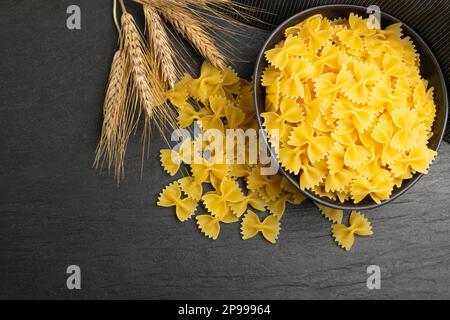 Rohe Farfalle Pasta in der Schüssel, gelbe trockene Nudeln mit Schmetterlingen, Makkaroni mit Weizenschleife, ungekochter Farfalle-Haufen auf schwarzem Hintergrund, Draufsicht Stockfoto