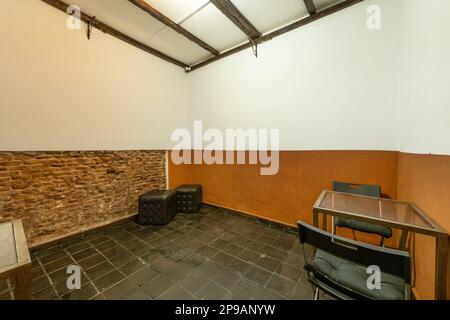 Ein fast leerer Raum mit schwarzen Fliesenböden, Ziegelwänden und einem Metalltisch mit zwei Klappstühlen und schwarzen quadratischen Puffs Stockfoto