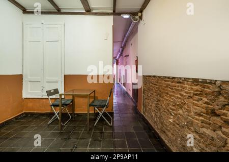 Ein fast leerer Raum mit schwarzen Fliesenböden, Ziegelwänden und einem Metalltisch mit zwei Klappstühlen Stockfoto