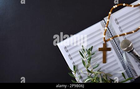 Hintergrund für Chormusik bei Palm Sunday mit Notenblättern, Mikrofon und Olive-Zweigen mit einem christlichen Kreuz auf einem schwarzen Tisch. Draufsicht. Stockfoto