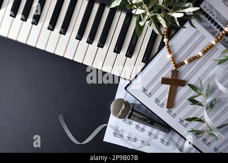 Religiöse Musik mit Klavier und Chöre auf einem schwarzen Tisch, dekoriert mit Olivenzweigen und einem christlichen Kreuz für die Feiertage des Palmensonntags. Draufsicht. Stockfoto