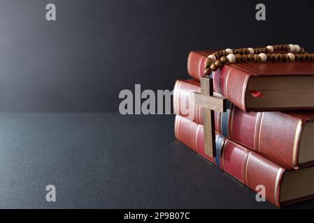 Hintergrund mit einem Stapel religiöser Bücher und religiösem Kreuz mit Kette aus Holzperlen auf schwarzem Tisch und Hintergrund Stockfoto