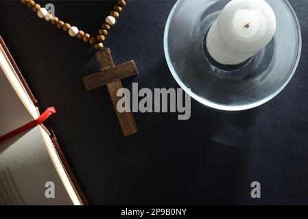 Schwarzer Nachttisch mit religiösen Elementen zum Beten mit einem christlichen Kreuz, einer offenen Bibel und einer beleuchteten Kerze auf einem Glasteller. Draufsicht. Stockfoto