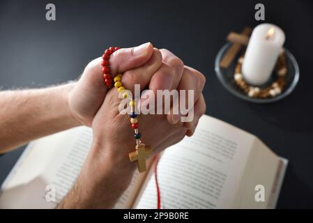 Christian betet mit Rosenkranz in den Händen, Daumen drücken und bibel und Kerze auf dem Tisch im Hintergrund Stockfoto