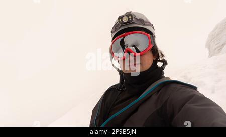 Kletterer mit Helm, Stirnlampe und schwarzer Jacke macht ein Selfie auf einem Gletscher an einem bewölkten Tag, die Landschaft spiegelt sich in rot gerahmten Gläsern wider Stockfoto