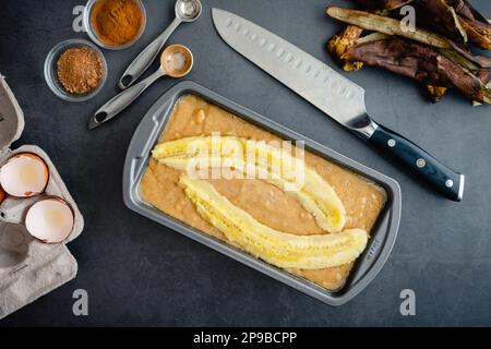 Bananenbrotteig in einer Brotpfanne und gebackener Teig: Ungebackener Teig in einer antihaftbeschichteten Brotpfanne, umgeben von Zutaten Stockfoto