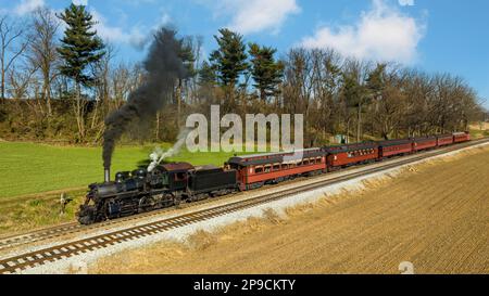Ein Blick auf die Drohne eines restaurierten Dampflokomotivzugs, der durch Farmland reist und an einem Herbsttag zu einem kleinen Bahnhof fährt Stockfoto