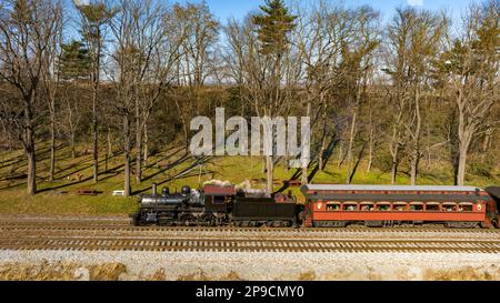 Ein Blick auf die Drohne eines restaurierten Dampflokomotivzugs, der durch Farmland reist und an einem Herbsttag zu einem kleinen Bahnhof fährt Stockfoto