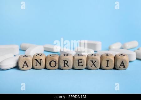 Word Anorexia aus Holzwürfeln mit Buchstaben neben den Pillen auf hellblauem Hintergrund Stockfoto