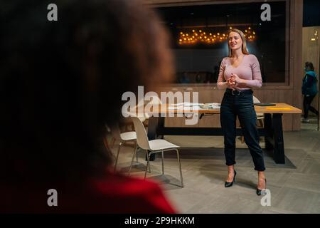 Blick von der Schulter einer nicht wiedererkennbaren Person bis hin zu einer jungen blonden Geschäftsfrau, die ein Treffen mit multiethnischen Kollegen leitet, im Büro interagiert. Stockfoto