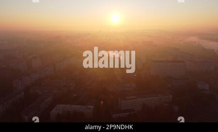 Wunderschöne Stadtlandschaft im Sommer bei Sonnenaufgang. Sie fliegen über viele mehrstöckige Gebäude und einen Schornstein mit weißem Rauch. Helle, glänzende Sonne. Stadtlandschaft. Sonnenaufgang Sonnenaufgang Sonnenaufgang Sonnenaufgang, aufgehende Sonne. Stockfoto