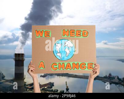 Der Demonstrante hält ein Plakat mit Text Wir brauchen eine Veränderung und eine verschwommene Sicht auf die industrielle Fabrik im Hintergrund. Klimaschutz Stockfoto