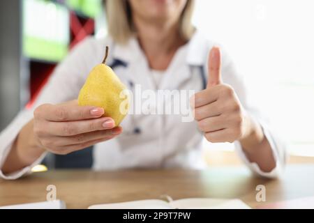 Ernährungsberaterin zeigt gelbe Birnen und Daumen hoch Geste, während sie am Kliniktisch sitzt. Stockfoto
