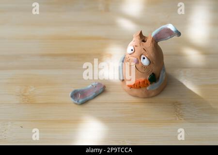 Clay Bunny mit gebrochenem Ohr hält Karotte, steht auf einem sonnigen Holzhintergrund. Top View Handmade Rabbit Universelles Symbol Für Ostern. Stockfoto