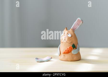 Sad Clay Rabbit Bunny mit gebrochenem Ohr hält Karotte und schaut weg. Handwerkskunst-Hase Universal Symbol Für Ostern. Stockfoto