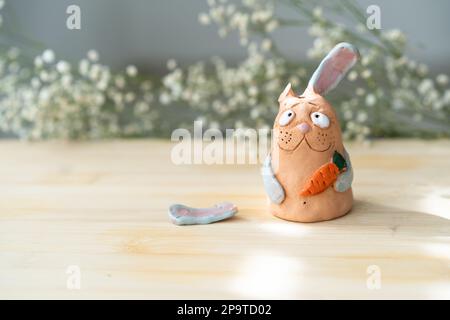 Clay Miniatur Hase mit gebrochenem Ohr hält Karotten auf wunderschönem Blumenhintergrund. Handgemachter trauriger Hase mit gebrochenem Ohr schaut auf die Kamera. Stockfoto