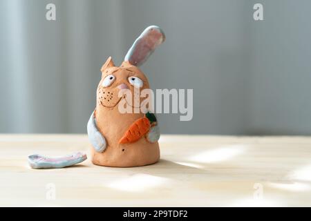 Sad Clay Rabbit mit gebrochenem Ohr hält Karotte und schaut in die Kamera. Handgemachtes „Bunny Universal“-Symbol Für Ostern. Stockfoto