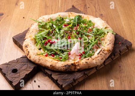 Im Ofen gekochte italienische Pizza Melagrano mit Mozzarella-Käse, Schinken, Rindfleisch, Rucola, Granatapfelsamen, basilikum und Olivenöl. Heiße knusprige Pizza auf einem Holzbett Stockfoto