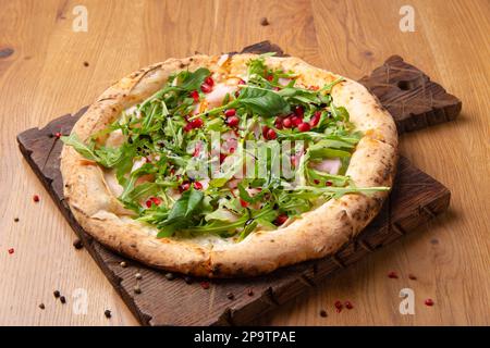 Im Ofen gekochte italienische Pizza Melagrano mit Mozzarella-Käse, Schinken, Rindfleisch, Rucola, Granatapfelsamen, basilikum und Olivenöl. Heiße knusprige Pizza auf einem Holzbett Stockfoto