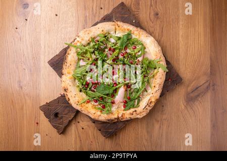 Im Ofen gekochte italienische Pizza Melagrano mit Mozzarella-Käse, Schinken, Rindfleisch, Rucola, Granatapfelsamen, basilikum und Olivenöl. Heiße knusprige Pizza auf einem Holzbett Stockfoto