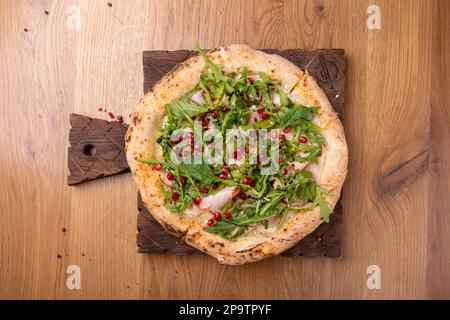 Im Ofen gekochte italienische Pizza Melagrano mit Mozzarella-Käse, Schinken, Rindfleisch, Rucola, Granatapfelsamen, basilikum und Olivenöl. Heiße knusprige Pizza auf einem Holzbett Stockfoto