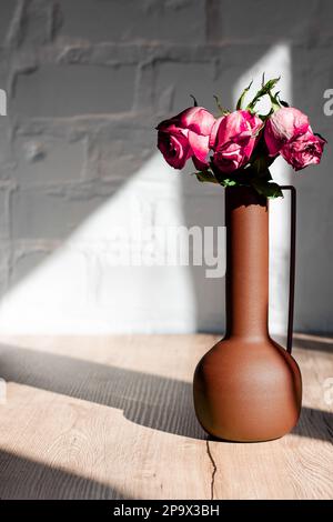 Verwelkte rosa Rosen in einer Vase in den harten Sonnenstrahlen auf einem Tisch vor einem Hintergrund aus weißen Ziegeln Stockfoto