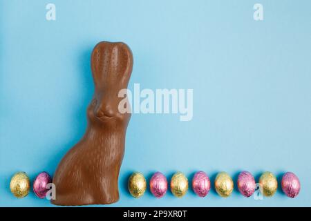 Milchschokoladenhasen und eine Reihe Osterbonbons, die in rosa und goldene Folie auf blauem Hintergrund verpackt sind. Frohes Osterkonzept. Vorbereitung auf den Urlaub. S Stockfoto