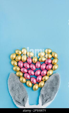 Osterhäschen-Ohren und herzförmiger Haufen Süßigkeiteneier, umhüllt mit rosa und goldener Folie auf blauem Hintergrund. Frohe Ostern, Liebeskonzept. Vorbereitung für Stockfoto