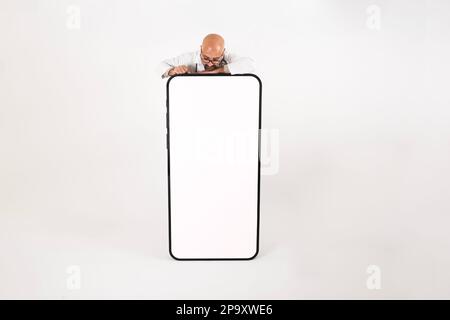 Ein Mann, der sich von hinten ein großes Smartphone-Modell anlehnt. Hellgrauer Hintergrund. Speicherplatz kopieren. Ein Typ, der auf dem weißen Bildschirm eines lebensgroßen Handys sitzt. Stockfoto