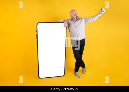 Lebensgroße ganze Körperlänge eines weißhäutigen Mannes, der Selfie mit einem riesigen Handy macht. Gelber Studiohintergrund mit Kopierbereich. Leerer leerer Bildschirm. Stockfoto