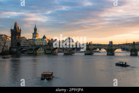 Blick auf den Sonnenuntergang über der Karlsbrücke in Prag, Fußgänger auf der Brücke, Bootsfahrten und Touristenattraktionen Stockfoto