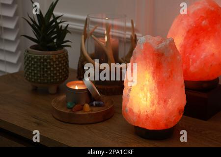 Salzlampen, Kerzen, Zimmerpflanzen und Edelsteine auf einem Holztisch in der Nähe weißer Innenwände Stockfoto