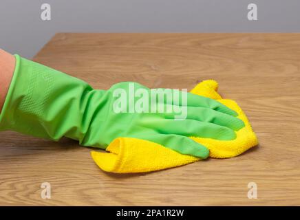 Grüner Handschuh zum Abwischen von Staub vom Holzschreibtisch, Reinigen des Holztisches mit einem Mikrofasertuch, gelber Wischer. Stockfoto