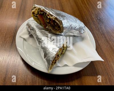 Weißer Teller mit köstlichem Schawarma, in Folie auf einem Holztisch verpackt. Köstliches nahöstliches Essen. Authentisches Street Food. Stockfoto