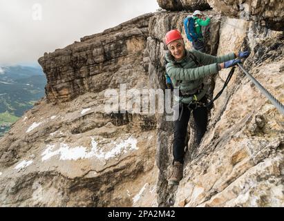 Gruppe junger Bergsteiger auf einer steilen Via Ferrata mit einem grandiosen Blick auf die italienischen Dolomiten in Alta Badia dahinter Stockfoto