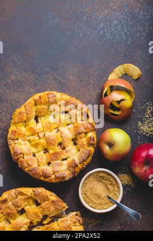 Blick von oben auf köstlichen hausgemachten Apfelkuchen mit geschnittenen Scheiben, frischen Äpfeln, Schale, Rohrzucker auf braunem rustikalem Steinhintergrund. Herbst oder Thanksgiving Stockfoto