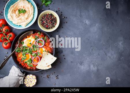 Shakshuka mit Pita-Brot in der Pfanne, Hummus in der Schüssel auf rustikalem Hintergrund. Traditionelle Gerichte aus dem Mittleren Osten. Spiegeleier mit Gemüse. Draufsicht. Leerzeichen Stockfoto