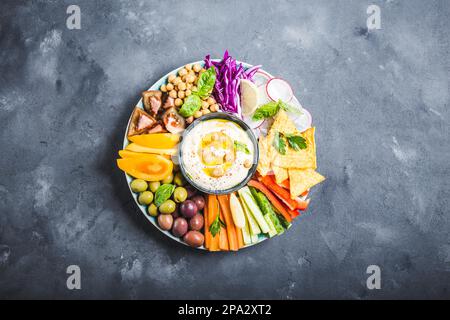 Hummus-Platte mit verschiedenen Snacks. Hummus in der Schüssel, Gemüsestangen, Kichererbsen, Oliven, Pita-Chips. Teller mit mediterranem Meze aus dem Mittleren Osten. Stockfoto