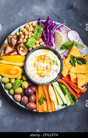 Hummus-Platte mit verschiedenen Snacks. Hummus in der Schüssel, Gemüsestangen, Kichererbsen, Oliven, Pita-Chips. Teller mit mediterranem Meze aus dem Mittleren Osten. Stockfoto