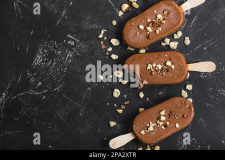 Milchschokolade Eis am Stiel mit Haselnüssen. Nahaufnahme. Eis am Stiel mit Schokolade, Stäbchen und schwarzem Steinhintergrund. Platz für Text. Oben Stockfoto