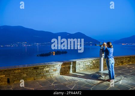 Frau, die in der Dämmerung ein Teleskop über dem Lago Maggiore mit den Bergen und den Brissago Inseln in Tessin, Schweiz, benutzt. Stockfoto