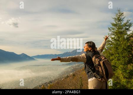Frau mit Rucksack und ausgestreckten Armen Genießen Sie einen Panoramablick über Locarno mit Wolkenlandschaft in Ticino, Schweiz. Stockfoto