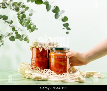 Probiotische Lebensmittel. Nachhaltiges Stillleben mit eingelegtem oder fermentiertem Gemüse mit der Hand einer Frau. Lecho und Ajvar in Glasgefäßen auf Netzbeutel auf blauem Backgr Stockfoto