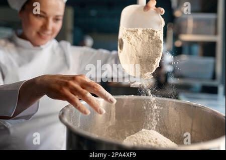 Weibliche Süßwarenfrau in weißer Uniform, die Mehl in eine große Metallschüssel schüttet Stockfoto