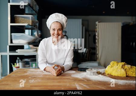 Porträt eines positiven Bäckers in Uniform, der mit Teig auf einen Holztisch geneigt wurde Stockfoto