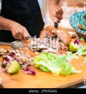 Mann, der Porchetta schneidet, ein italienisches Gericht, entbeintes Schweinerollchen, gefüllt mit einer Mischung aus Kräutern, Gewürzen und Knoblauch. Stockfoto