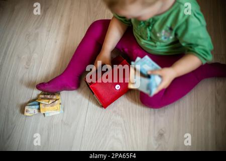 Mädchen Kleinkind mit roter Handtasche und Banknoten in den Händen sitzt im Zimmer auf dem Boden. Das Kind spielt mit Geld. Finanzkrise. Leere Handtasche. Stockfoto