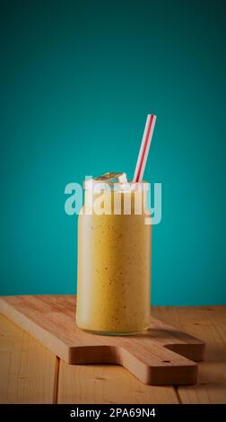 Frucht-Smoothie-Getränk in einem Glasgefäß mit Eis und einem Strohhalm, auf einer Holzfläche mit blauem verlaufendem Hintergrund. Stockfoto